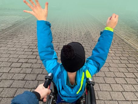 Eine Person im Rollstuhl hebt im Freien beide Arme.